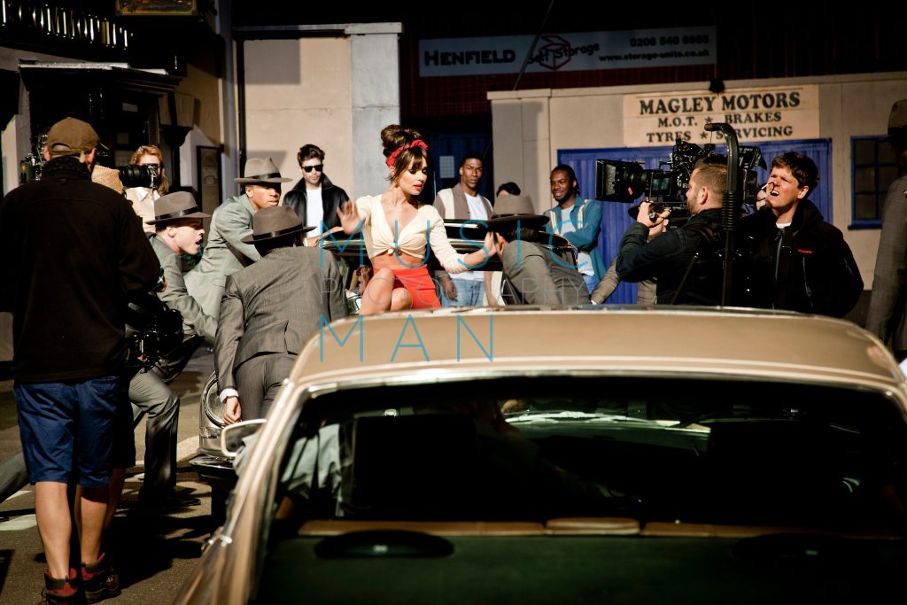 Singer Cheryl Cole performing on a classic car during a high-budget music video production set for Under the Sun at Wimbledon Studios, featuring film crews, cameras, and background extras, behind-the-scenes photography by Music Man Studio.