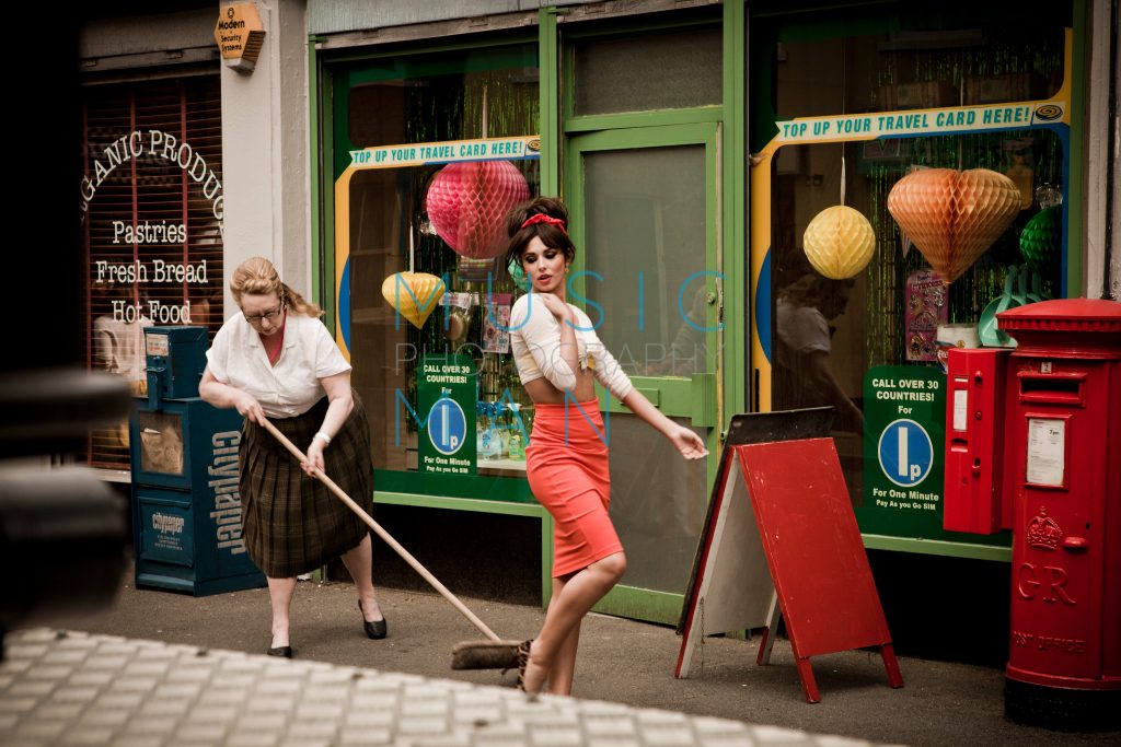 Pop singer Cheryl Cole in a coral pencil skirt posing next to a vintage corner shop front and a woman sweeping the street on the Under the Sun music video set at Wimbledon Studios, photography by Music Man Studio.
