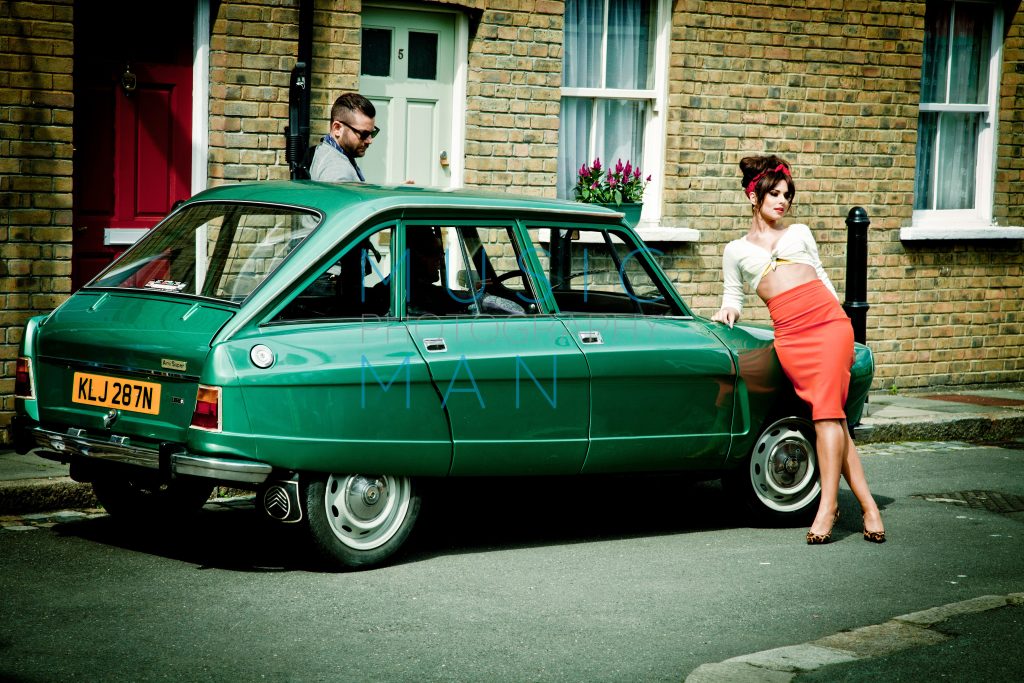 Singer Cheryl Cole in a retro coral pencil skirt posing against a classic green vintage Citroën Ami Super car on a 1960s backlot street set at Wimbledon Studios, behind-the-scenes photography by Music Man Studio.