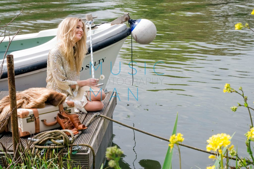 Singer Diana Vickers in a gold embellished dress sitting on a wooden river dock next to a vintage suitcase and a white boat named Salty, music video photography by Music Man Studio.